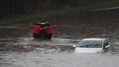 An elderly man is helped out of a car in waist-deep floodwater by two members of the fire service