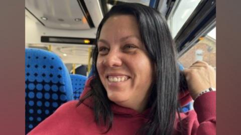 Agata Wiznefska is smiling. She has long, dark hair and is wearing a red hoodie. She appears to be sat on a bus or train seat.