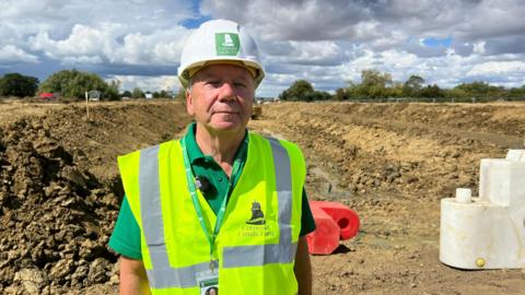David Hagg stands in front of a canal channel, which has no water in it and is just a trench of mud. It is a sunny day and he wears a green T-shirt underneath a hi-vis vest. He has a white hard hat on.