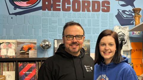 Stephen Thompson and his partner Rebecca. They are standing side-by-side in front of a sign that says Regency Records. Stephen is wearing a black hoodie with a small logo on the left side of the chest that says Record Store Day, and Rebecca is wearing a blue hoodie that has a logo in the centre saying Regency Records. Stephen has black hair, and is wearing glasses. Rebecca has dark brown hair.