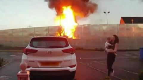 A fire takes hold in a fence, as a woman carrying a baby runs to a white car parked in a car park, opposite the fire