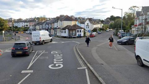 Google Street View image of the junction of Grove Place and Avenue Road in Falmouth. A white van and black VW car are travelling along Grove Place. Two pedestrians are walking on the pavement on the right-hand side of the road.