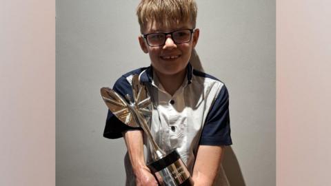 Luke Mortimer is pictured against a grey background wearing a black and white shirt and holding his large Pride of Britain award in the shape of a silver winged woman.