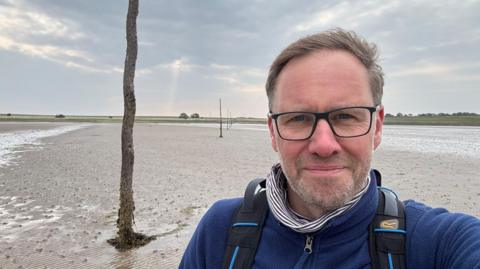 Gareth Davies-Jones standing on a beach doing a selfie. He has black glasses and short brown hair. Here is a white stubble. He is wearing a blue jacket and a backpack. 