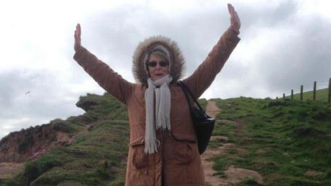 A woman in her seventies is pictured with her arms in the air. She is wearing a brown coat with a fur-lined hood pulled up over her head and a grey scarf. She is standing on a hill with a path going uphill behind her.