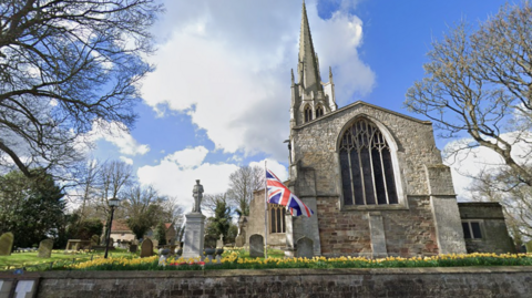 A church with a large stained glass window on the gable end and a spire, set within a graveyard. There is a row of daffodils, a union flag and a war memorial in the graveyard. Trees in the background and a blue sky.