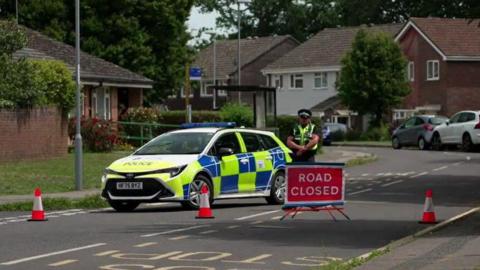 Police officer standing behind cordon on a road with police car behind and homes in the background.