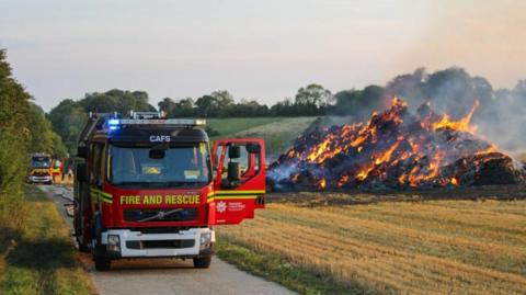 Two fire engines on a track with a large pile of hay on fire in a field to the right.