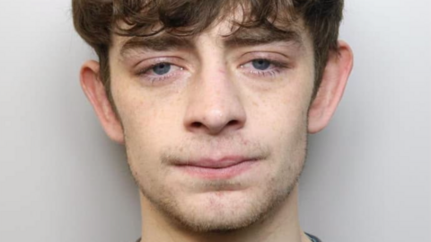 Mugshot of Ronnie. He has stuble around his chin. Floppy brown hair which covers his thick eyebrows. He is standing against a grey backdrop.