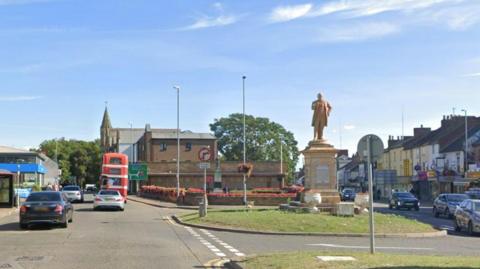 Abington Square, Northampton, showing statue of a man on a plinth (Charles Bradlaugh), which is standing on a grass island. To the left is a blue canopy of a car showroom or similar, there is a church in the centre of the picture and shops with flats above to the right. There are some cars and a red double decker bus on the road to the left.