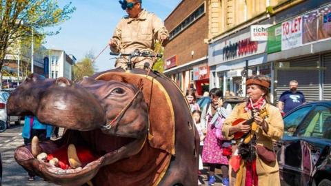 A large mechanical hippo is being ridden through a town centre street by a man wearing strange goggles. A woman is standing alongside playing a musical instrument and there are shops behind.