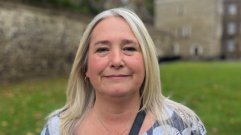 Claire is wearing a patterned top, she has long blonde hair and is standing on College Green, Westminster, being interviewed.