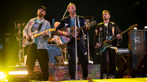 Coldplay on stage with Chris Martin up front and a lead guitarist to the left and bass player to the right. There is also a drummer in the background and yellow lighting. 