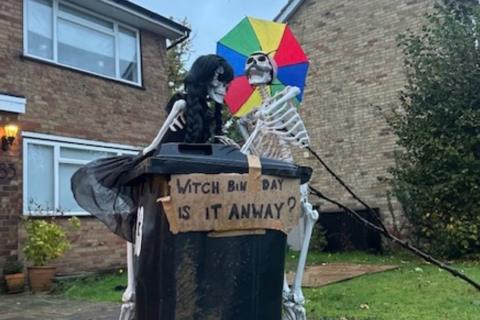 Two skeletons stood by a bin, one is holding an umbrella. There is a sign that says 'Witch bin day is it anyway?'