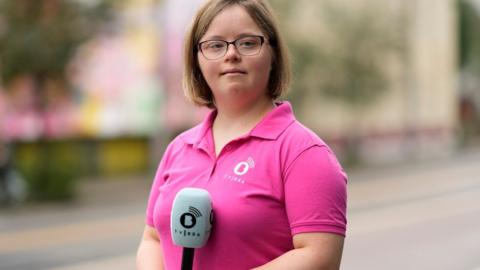 A young woman with Down's syndrome stands in a street, holding a microphone. She is dressed in a bright pink polo shirt. Her hair is medium brown and not quite shoulder length. She is wearing glasses and has a hint of a smile. 