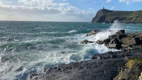 A seascape with waves crashing on the rocky shore, blue sky, white clouds and a cliff in the background