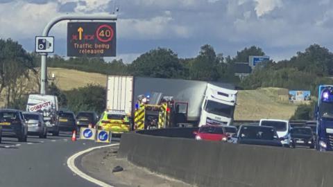 A jackknifed lorry lies across numerous lanes of a motorway. Traffic can be seen on each side as well as emergency services.