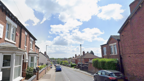 A Google street view screen shot of a residential road. A range of terraced and semi-detached homes can be seen from the road junction. Cars are parked on the road