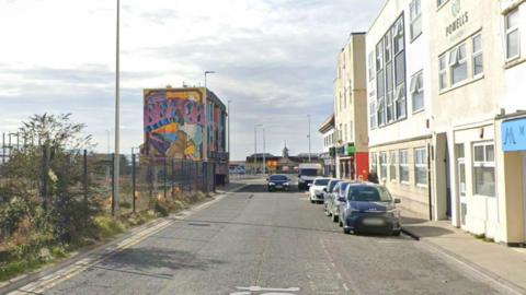 A Google maps street view of Oxford Street in Weston-super-Mare. There are cars parked on the side of the road and there is a building covered in graffiti in the distance. A row of buildings can be seen on the opposite site of the road.