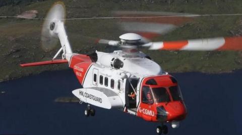 A red and white HM Coastguard helicopter in flight over some coastline.