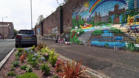 A new garden planted on a road next to a pavement and parked car. A brightly coloured mural depicting the Liverpool skyline is painted on a wall next to the pavement.