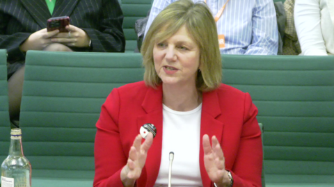 Minister Lilian Greenwood gives evidence to the transport committee, wearing a white scoop-necked t-shirt, a scarlet jacket and a wristwatch. 