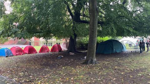 Eight tents and gazebos can be seen erected under two trees in Weston Park. Three people can be seen on the right with one of them standing in front of a shopping trolley.