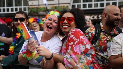 A picture focused on two attendees of a previous Pride in London event. On the left, a woman with glitter on her face and with flowery decorations around her head and arms laughs at the camera. On the right, another woman smiles at the camera with red-heart sunglasses on and a red flowery dress.