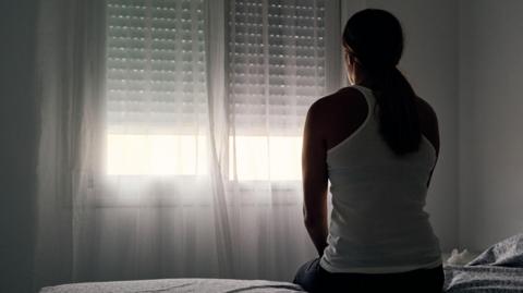 A woman with a long dark pony tail sits on a bed facing a window with the curtains and blind closed. She is wearing a white vest top. There is daylight coming through the lower part of the window and the walls are white.