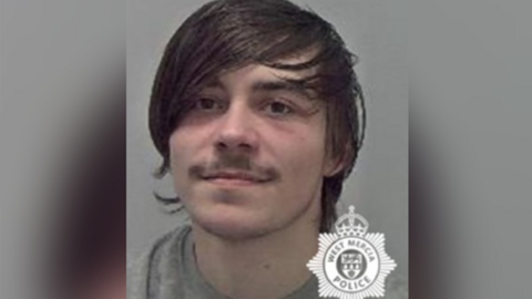 A mug shot of a man with long brown hair and a brown moustache, wearing a grey sweater