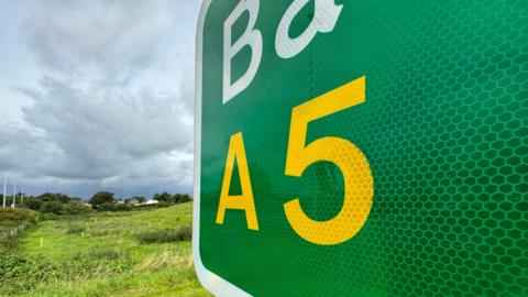 Close-up of green UK road sign displaying A5 with green fields and farmhouse in the background