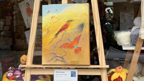 A painting of red and blue birds on an easel is shown in the window of a business. 