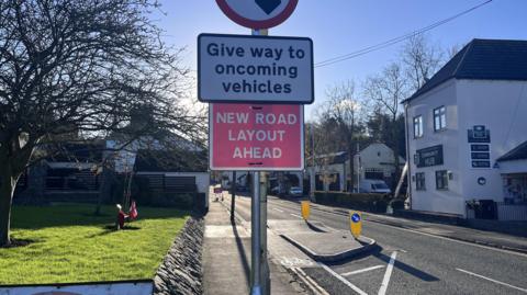 A build-out on the left side of the road which makes the street only one lane wide. A sign prominently in the photo reads "Give way to oncoming vehicles" and a sign below says "new road layout ahead"