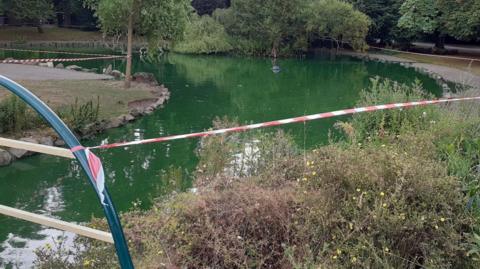 A close up shot of a park with a spherical pond containing green water. There is an island in the middle and bushes in front and behind the pond with a white and red tape surrounding it.