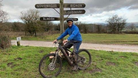 Laurence Penn is wearing a blue coat and cycling helmet. He is sitting on his bicycle on a field. Behind him are road signs. 