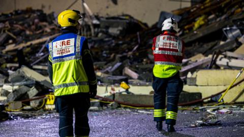 Two firefighters are standing in front of a large pile of rubbish on the site of the explosion. They are in uniform and are wearing hard hats. The man standing closest to the camera has FIRE SAFETY OFFICER printed on the back of his jacket.