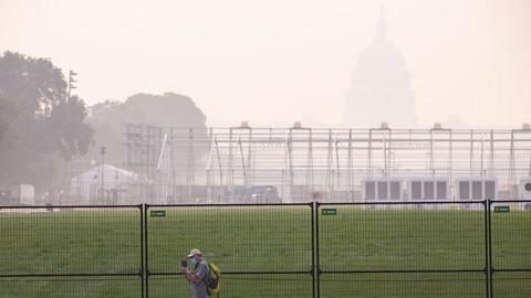Man walks in front of fencing and US Capitol amid thick haze, wearing face mask