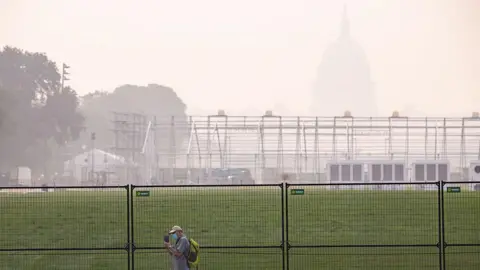 Man walks in front of fencing and US Capitol amid thick haze, wearing face mask