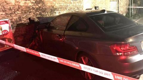 A grey BMW car has collided with an old stone wall. There is red and white cordon tape in the foreground.