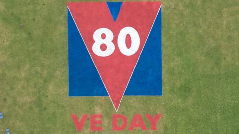 A red and blue logo reads '80 VE DAY'. It is sprayed onto green grass.