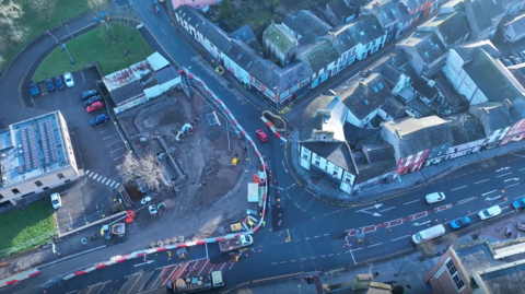 An aerial view of roadworks in Workington town. There are red and white striped barriers around a large construction area on the left of the image and traffic cones separating the road lanes.