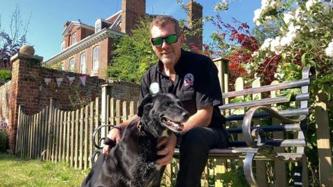 A man wearing a black shirt and sunglasses with a black dog in front of him. They are in a garden beside a house on a sunny day