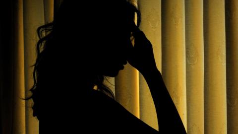 The silhouette of a woman with her hand on her forehead, distressed, with yellow blinds behind her.