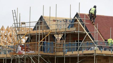 Construction site shows roofs of houses under construction. The houses are surrounded by scaffolding and two workers wearing high-visibility jackets are working on the roof. 
