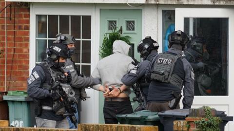 A man in a grey hoodie facing away from the camera, with handcuffs on behind his back. He has armed police officers either side of him, with the letters ‘CTSFO', 'EMOE' and 'POLICE' on their uniforms.