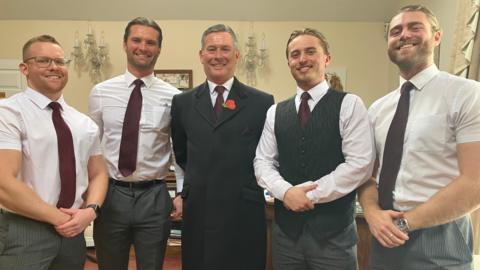 Five men wearing white shirts and red ties stand in a line, smiling at the camera. The middle one is wearing a long black coat and a red poppy, and another has a black waistcoat.