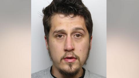 A man with dark hair and a goatee beard wearing a police issue grey sweatshirt