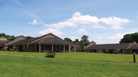 The picture shows a wide outdoor view of a low, spread‑out brown building complex set in a large grassy area. The buildings appear to be single storey, with pitched roofs covered in dark tiles and light‑coloured walls. 