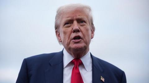 Donald Trump speaking outdoors against a pale, cloudy sky. He is wearing a dark navy suit, a white dress shirt, and a bright red tie, with a small American flag pin on his lapel.