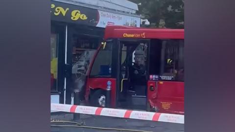 A red bus can be seen partway through a smashed window at the front of a store. There is red and white tape to identify a cordon around the scene.